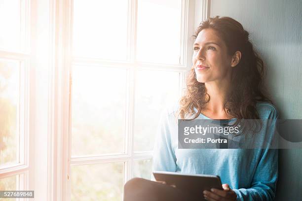 pensierosa giovane donna che tiene compressa digitale dalla finestra - sognare ad occhi aperti foto e immagini stock