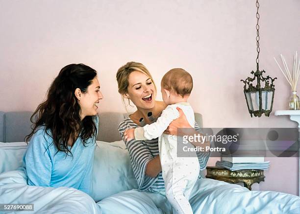 happy young lesbians playing with baby boy in bed - lesbische stockfoto's en -beelden