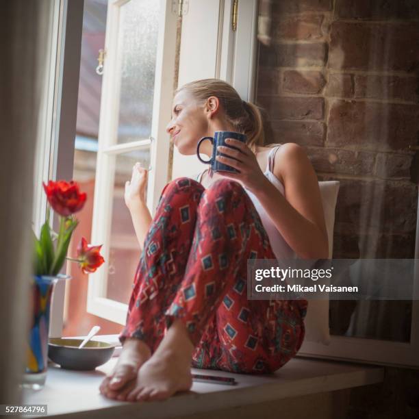 young woman sitting in pyjamas on windowsill - junge frau allein stock-fotos und bilder