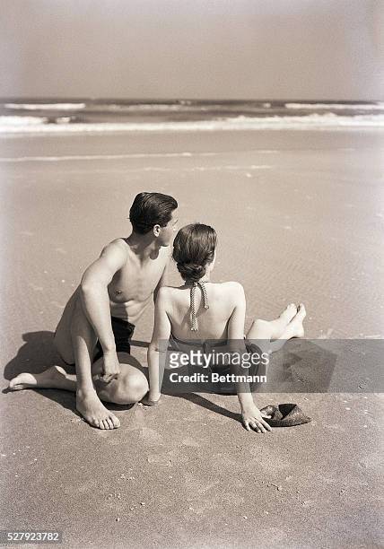 Photo shows a girl and a boy seated on a beach. Models: Mary Elizabeth Thomson and Sol Mendelson. Ca. 1940s-1950s.