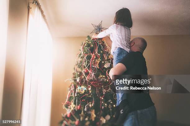 father helping daughter put star on christmas tree - leanincollection father fotografías e imágenes de stock
