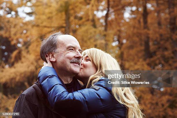 portrait of a mid-aged woman kissing her senior father on the cheek in a park - hija fotografías e imágenes de stock