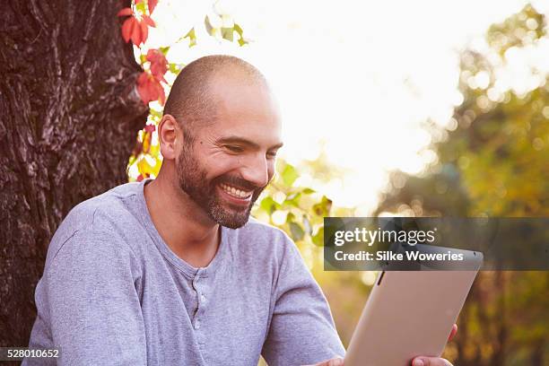 portrait of an adult man leaning against a tree trunk and working on his tablet computer, backlit. - mature man using phone tablet stock pictures, royalty-free photos & images