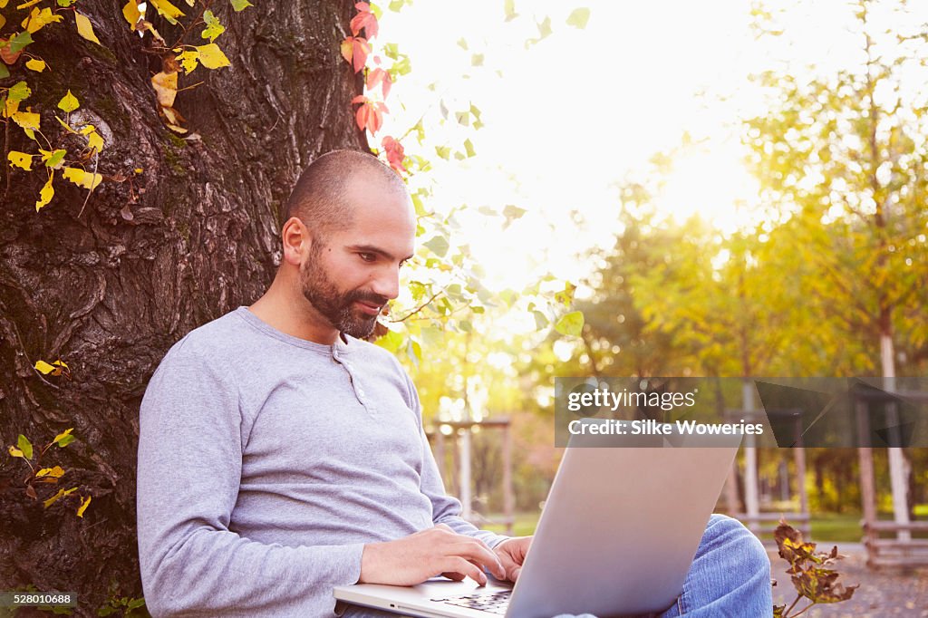 Portrait of an adult man leaning against a tree trunk and working on his laptop, backlit.