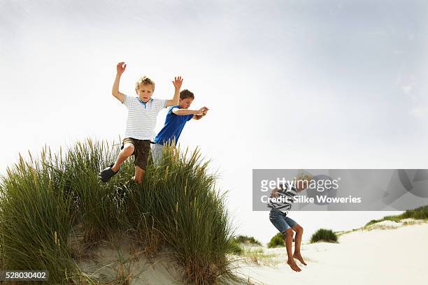 three boys jumping over a small dune on a beach - marram grass stock pictures, royalty-free photos & images