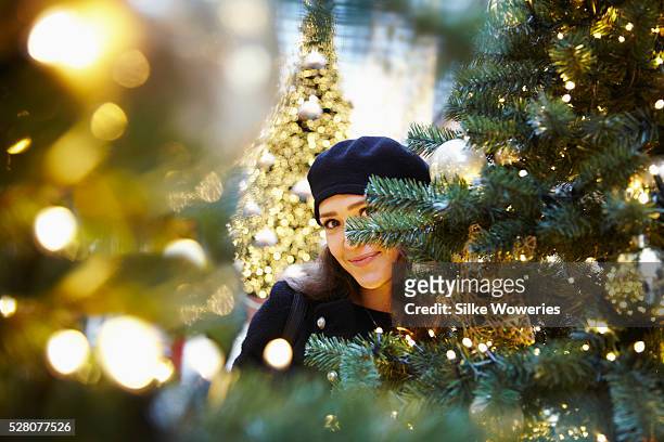 portrait of a young woman hiding between decorated christmas trees looking into the camera - decorated christmas trees outside stockfoto's en -beelden