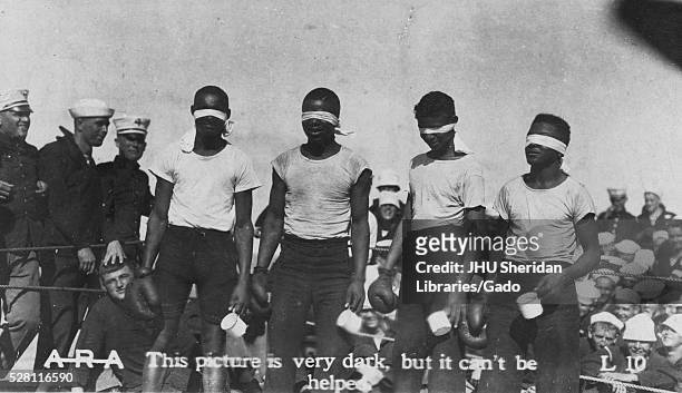 Four African American boys are standing in a boxing ring with blindfolds on, each has a boxing glove in one hand and a mug in the other hand, they...