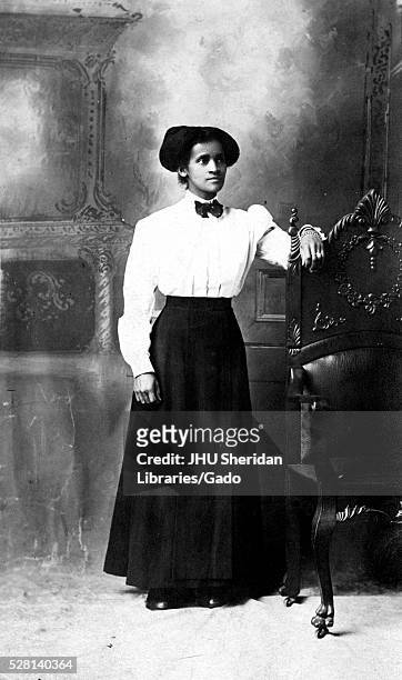 Full-length portrait of an African American woman standing in front of a decorative mural, she is resting one arm on a high wooden back of a chair,...