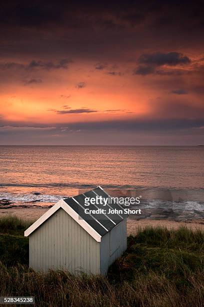 a small building along the water with a pink sky at sunset; bamburgh northumberland england - northumberland stock pictures, royalty-free photos & images