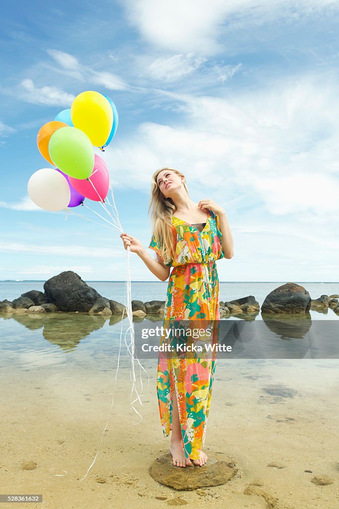 A young woman in a floral dress standing on the beach holding a bundle of colourful balloons; Kauai, Hawaii, United States of America