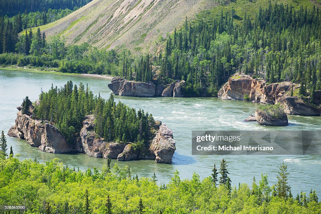 A small island covered with trees in a river with a steep slope of a mountain along the shoreline; Yukon, Canada