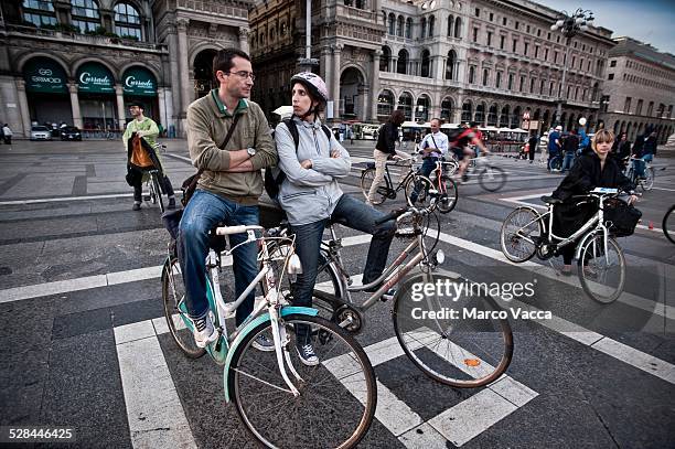Young couple sits on their bicycle and chat. Other cyclists stand by around Duomo square. Milano