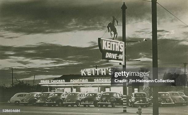 Black and white photographic postcard of Keith's Drive-In at night with cars parked in front.