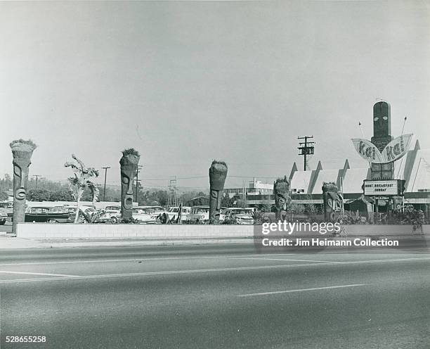 Black and white photograph of Polynesian style restaurant and parking lot with statues and cars.