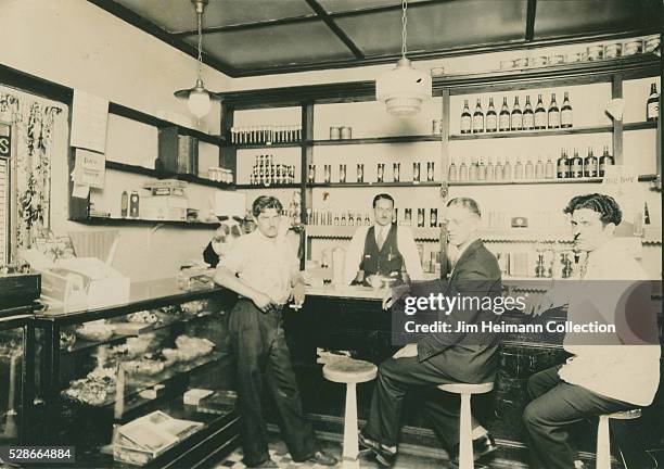 Black and white photograph of three men posing at bar and bartender behind bar. Wall in back lined with bottles.