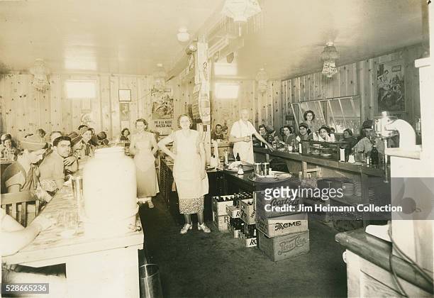 Black and white photograph featuring waitresses and diners at counter in restaurant.