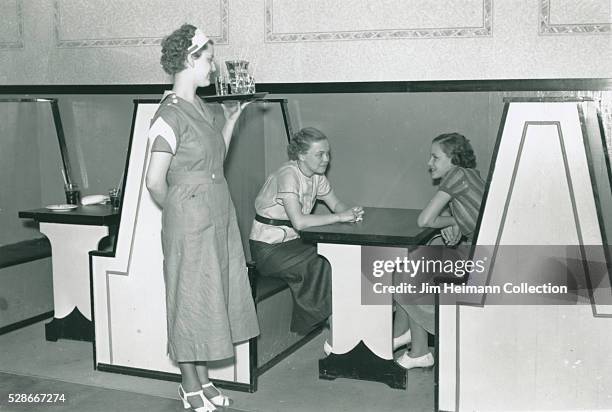 Black and white photograph of waitress holding tray and serving young women sitting in booth.