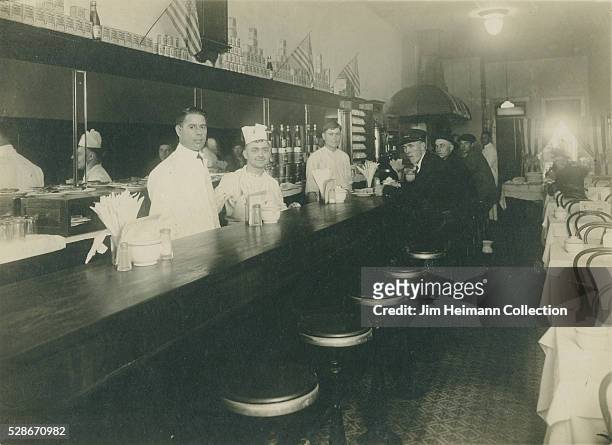 Black and white photograph featuring restaurant workers behind counter and patrons sitting at counter. American flags on wall.