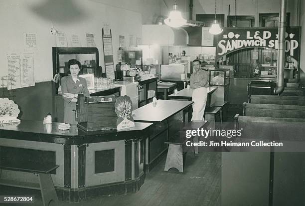 Black and white photograph featuring one man and one woman standing by cash register and tables.