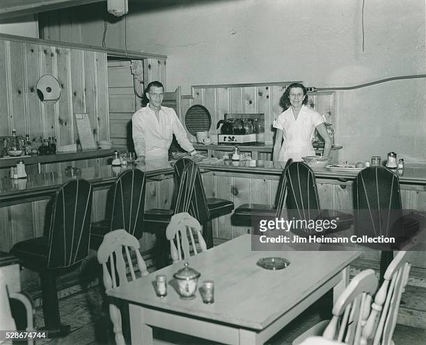 Black and white photograph of man and woman standing behind counter in wood paneled restaurant.