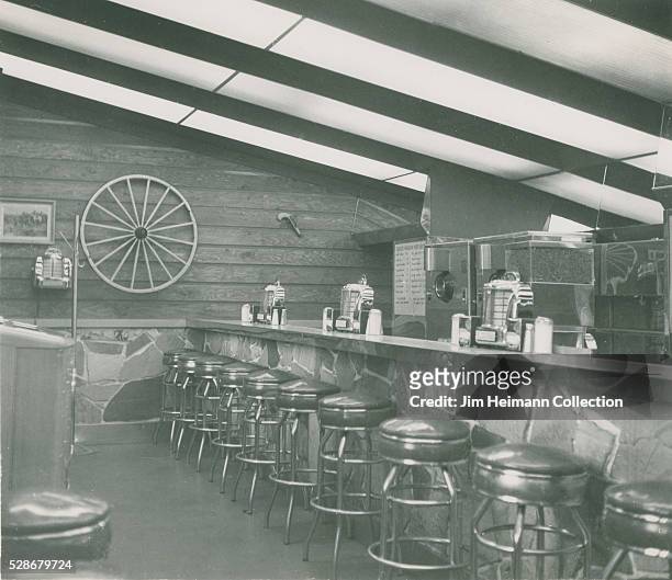 Black and white photograph of counter and stools in Chuck Wagon restaurant, with wagon wheel on wall and juke box.