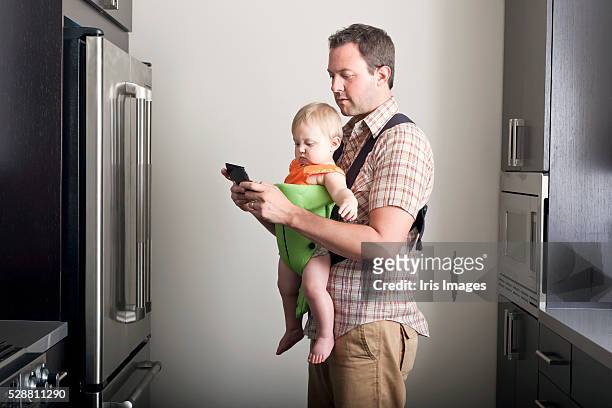 father carrying daughter (6-12 months) in carrier and text messaging - babytrage stock-fotos und bilder