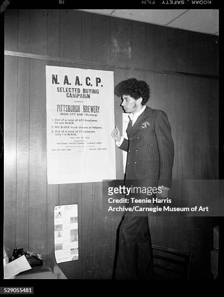 Tim Stevens posing on chair beside sign inscribed 'NAACP Selected Buying Campaign Against Pittsburgh Brewery,' in NAACP office, Pittsburgh,...
