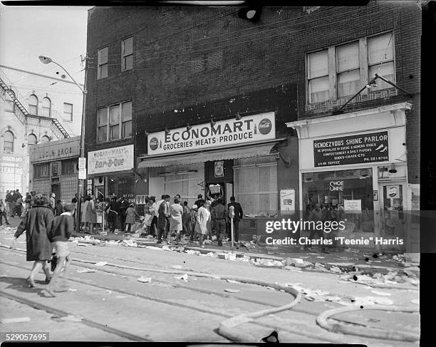 Men and women gathered outside Economart Market with broken windows, Rendezvous Shine Parlor, and Hogan and Mary's Bar-B-Q, with fire hoses in...