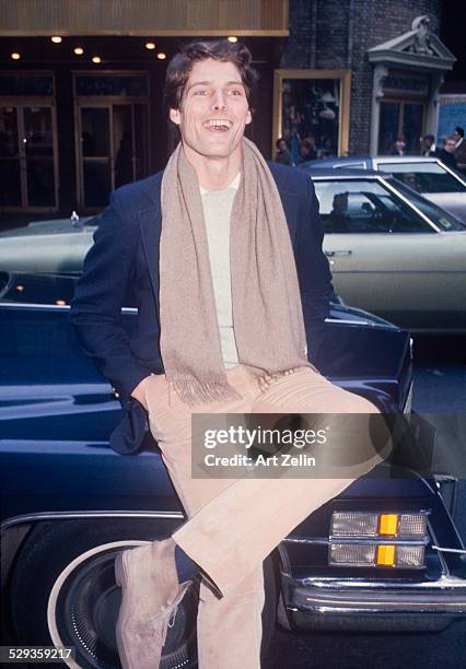 Christopher Reeve sitting on a car fender, laughing. Theater in the background; circa 1970; New York.