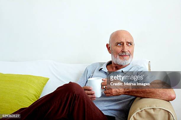 thoughtful senior man holding coffee mug on sofa - three quarter length stock pictures, royalty-free photos & images