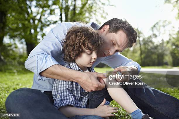 father applying bandaid to son's (7-9) knee in park - machucado imagens e fotografias de stock