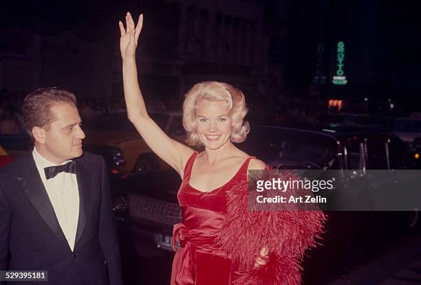 Carroll Baker with her husband Jack Garfein on the street. She is waving wearing a red dress and feather boa limousine in the background ; circa...