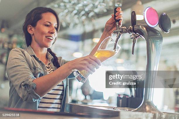 casual young cheerful female bartender pouring beer from facet - beer pump stock pictures, royalty-free photos & images