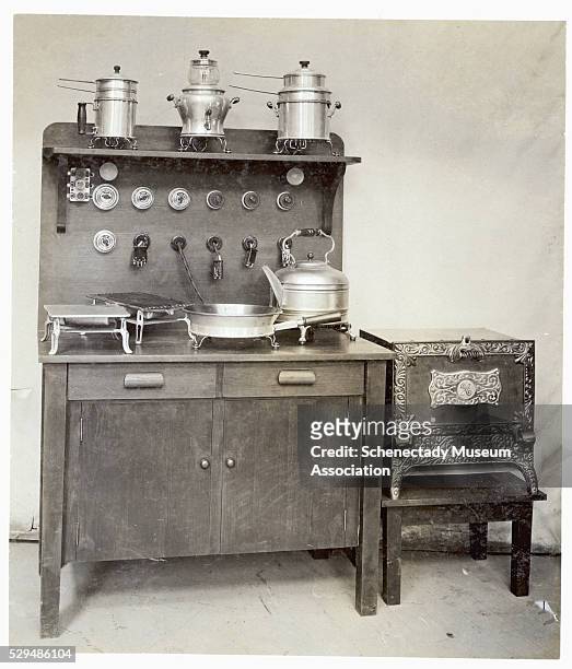 Cooking and baking table for electrical appliances has plugs running from it. A frying pan, kettle, and gridles sit on it, with pots above.
