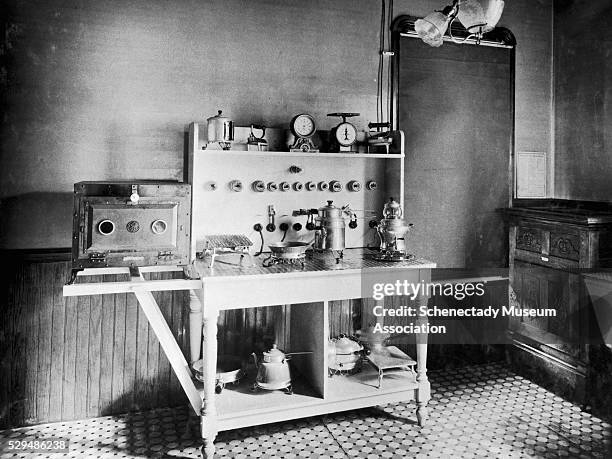 Cooking and baking table for electrical appliances has plugs running from it. A frying pan, kettle, and griddles sit on it, with pots below and...