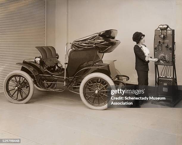 Young woman uses a hand-cranked battery charger to charge her electric Columbia Mark 68 Victoria automobile. The Pope Manufacturing Company of...