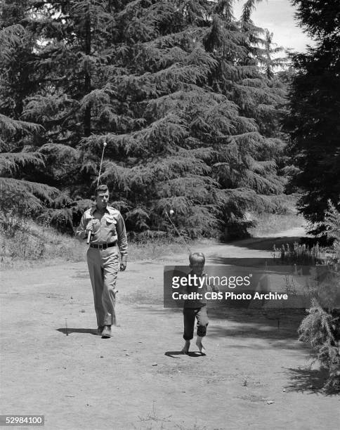 American actors Andy Griffith and Ron Howard walk along a dirt road with fishing poles over their shoulders in a scene from 'The Andy Griffith Show,'...