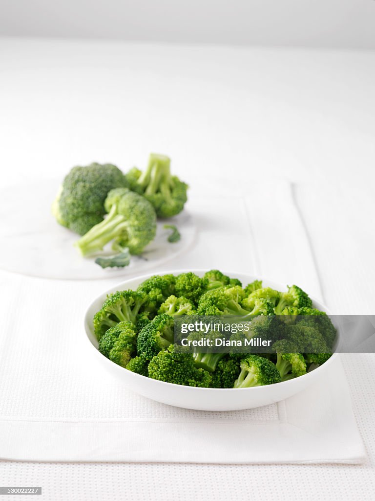 Raw broccoli on marble cutting board and bowl of boiled broccoli