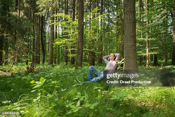 mature man leaning by tree in forest, smiling - leaning tree stock pictures, royalty-free photos & images