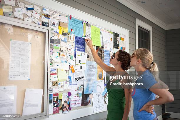 two mid adult women looking up at community notice board - bulletin stock pictures, royalty-free photos & images