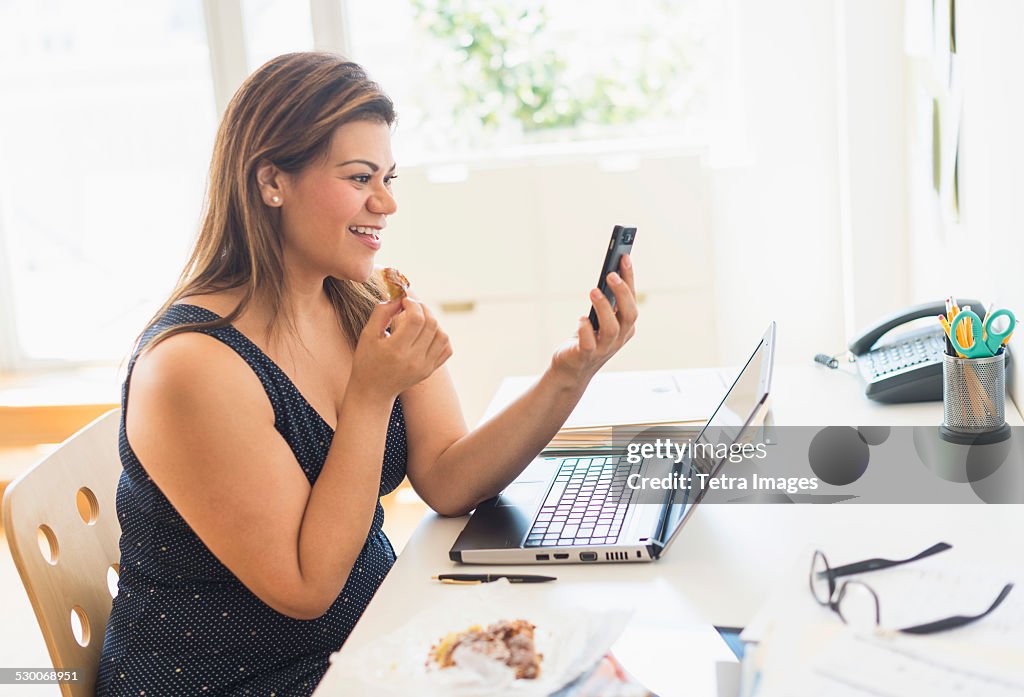 USA, New Jersey, Jersey City, Woman eating croissant and using mobile phone in office