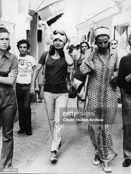 American first lady Jacqueline Kennedy Onassis wearing a kerchief on her head walks through a busy street with the Gaines girls in Capri, Italy,...