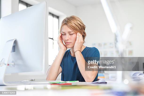 portrait of young woman with headache at her desk in a creative office - headache stock-fotos und bilder