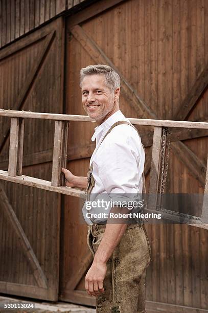 man in lederhosen carrying ladder on farm, barvaria, germany - bavarian man in front of house stock-fotos und bilder