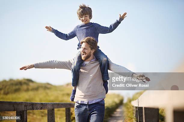 father carrying son on shoulders at the coast - llevar al hombro fotografías e imágenes de stock
