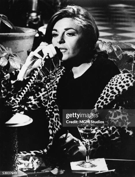 American actress Anne Bancroft wears a leopard print coat as she talks on the phone, drinks, and smokes in character as the seductive older woman,...