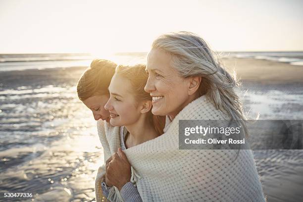 grandmother, mother and daughter wrapped in a blanket on the beach - drei frauen stock-fotos und bilder