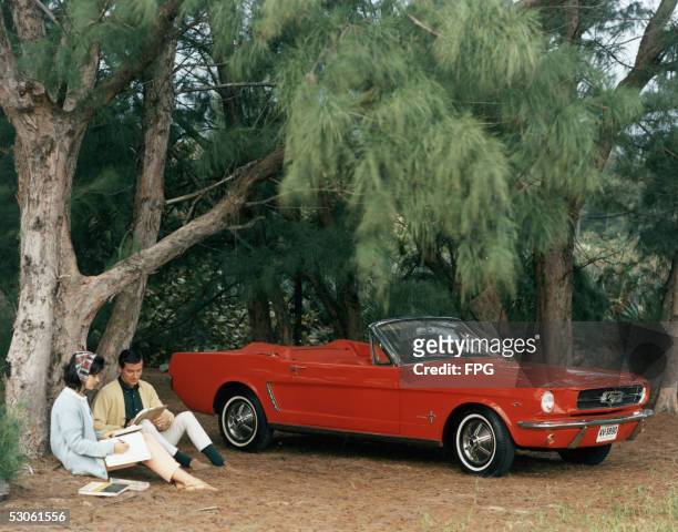 Promotional shot of a red 1964 Ford Mustang convertible parked in a forest clearing with a couple sitting by a pinetree, 1964. This first series of...