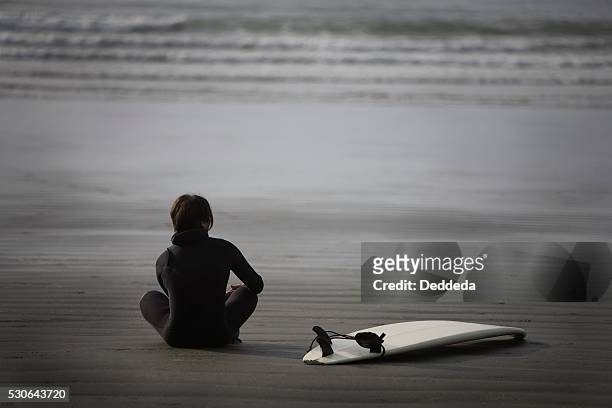 surfer sitting on beach, cox bay near tofino, british columbia, canada - surfer wetsuit crossed foto e immagini stock