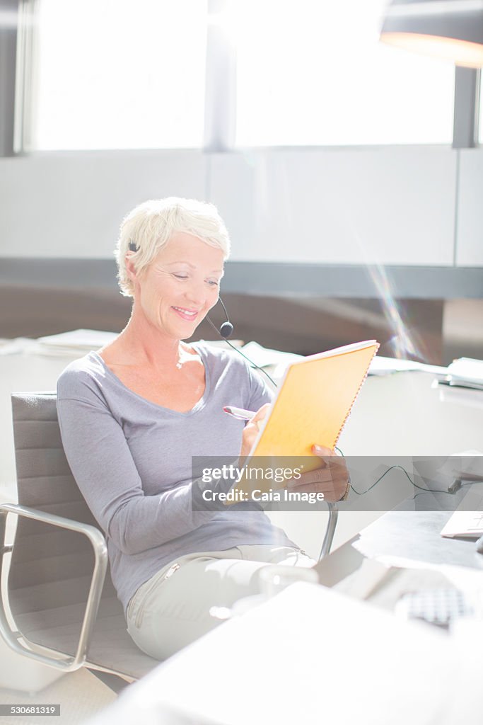 Businesswoman in headset writing in notebook at office desk